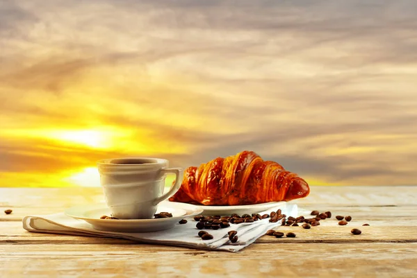 木製のテーブルの上にクロワッサンとコーヒー 日の出の背景に朝食 コーヒーとセラミックマグカップ マグカップの近くにコーヒー豆が散乱しています 食品の近くにテキストのためのスペースとフリースペースをコピー — ストック写真
