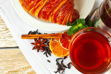 Beyaz tabakta kruvasan ve çay. Eski ahşap bir masada çay fincanı. Tam siyah çay, limon, nane yaprağı ve tarçın peçetede..