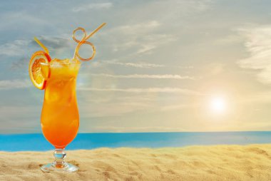 Plajda portakal kokteyli. Buzlu alkollü içecek, kumda portakal rengi. Arka planda gün batımı, deniz ve gökyüzü. Cama yakın metin için boşluk ve boş alanı kopyala.