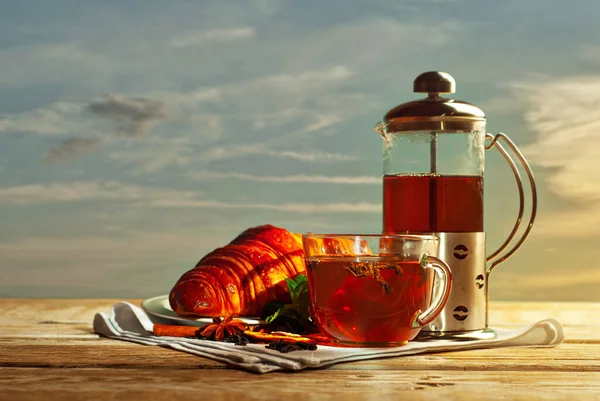 木製のテーブルの上にお茶とガラスティーポットのマグカップ 日没の背景にお茶とクロワッサン 白いナプキンに粗い紅茶 レモン シナモンとミントの葉 — ストック写真