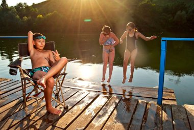 Çocuk gölün yanındaki bir sandalyede oturuyor. Çocuklar iskelede dinleniyor. Çocuklar suya atlıyor. Yaz, tatil, seyahat ve tatil kavramı.