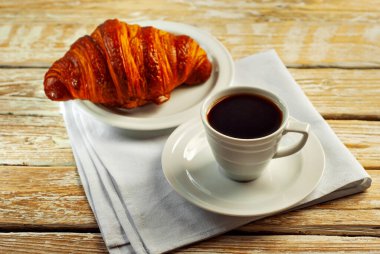 Beyaz seramik bardakta kahve. Kahvaltıda kruvasan ve kahve. Eski ahşap bir masada kahve..