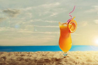 Plajda portakal kokteyli. Buzlu alkollü içecek, kumda portakal rengi. Arka planda gün batımı, deniz ve gökyüzü. Cama yakın metin için boşluk ve boş alanı kopyala.