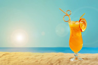 Plajda portakal kokteyli. Buzlu alkollü içecek, kumda portakal rengi. Arka planda gün batımı, deniz ve gökyüzü. Cama yakın metin için boşluk ve boş alanı kopyala.