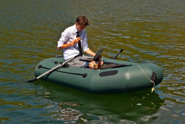 Şişme teknede beyaz gömlekli bir adam. Göl kenarında dizüstü bilgisayarı olan bir iş adamı. Uzaktan çalışma, boş zaman ve serbest çalışma kavramı..