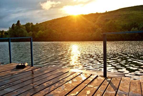 Пейзаж с озером и пирсом. Вода сверкает на фоне заката. Деревянный пирс на переднем плане.