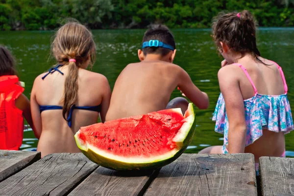 Children eat watermelon on the pier. Children in life jackets rest on the river bank. Camping by the water. Sharpness on watermelon.