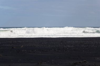 Dalgalar, ufuk çizgisi ve mavi gökyüzü üzerindeki siyah kumsal manzarası.