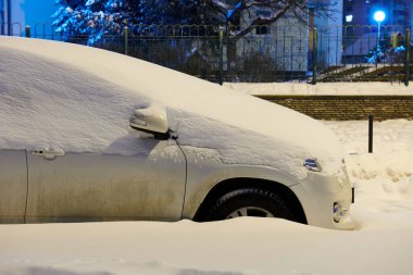 Geceleri karla kaplı beyaz bir araba. Bahçeye park edilmiş bir araba. Şehir üstüne kar fırtınası. Yan görüş. Yakın çekim..