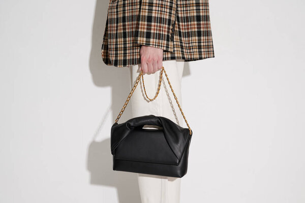 Женщина в клетчатом пиджаке и белых кожаных брюках с черной кожаной сумочкой с золотой цепью в руке. Белый фон. Горизонтальное фото.