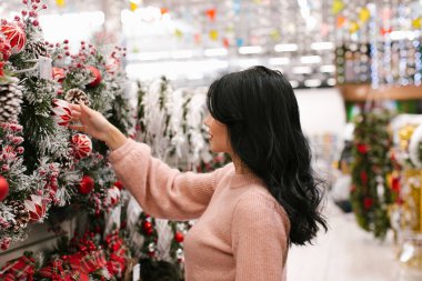 Pembe kazaklı genç esmer kadın mağazada bir Noel çelengi seçiyor. Yeni yıl alışverişi.