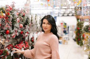Pembe kazaklı genç esmer kadın mağazada bir Noel çelengi seçiyor. Yeni yıl alışverişi.