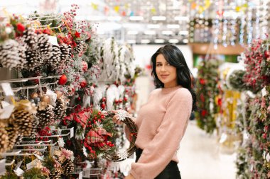 Pembe kazaklı genç esmer kadın mağazada bir Noel çelengi seçiyor. Yeni yıl alışverişi.