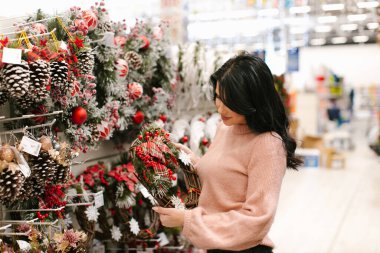 Pembe kazaklı genç esmer kadın mağazada bir Noel çelengi seçiyor. Yeni yıl alışverişi.