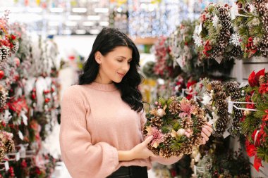 Pembe kazaklı genç esmer kadın mağazada bir Noel çelengi seçiyor. Yeni yıl alışverişi.