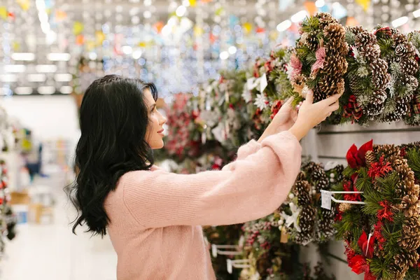 Pembe kazaklı genç esmer kadın mağazada bir Noel çelengi seçiyor. Yeni yıl alışverişi.