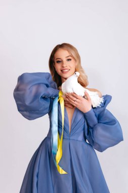 Ukrayna için dua et. Mavi kabarık elbiseli, Ukrayna bayrağı renginde kurdeleli genç bir kadın beyaz arka planda barışın beyaz güvercin sembolünü taşıyor. Savaşı durdurun. Boşluğu kopyala.