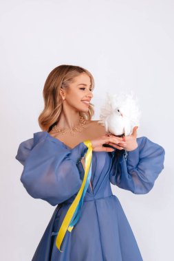 Ukrayna için dua et. Mavi kabarık elbiseli, Ukrayna bayrağı renginde kurdeleli genç bir kadın beyaz arka planda barışın beyaz güvercin sembolünü taşıyor. Savaşı durdurun. Boşluğu kopyala.