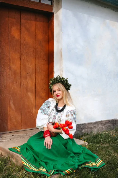 A Ukrainian woman in a traditional Ukrainian embroidered dress, with a wreath on her head, sits on the threshold of a house with a jar of red tulips in her hands. Slavic girl.