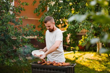 Beyaz tişörtlü, sakallı genç bir adam evinin yakınındaki bahçedeki ızgarada barbekü yapıyor. Adam ızgarada domuz kebabı kızartıyor..