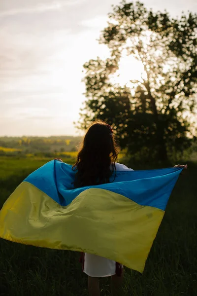 A Ukrainian woman in an embroidered Ukrainian traditional dress stands with her back in a field with a large flag of Ukraine in the field. Pray for Ukraine.