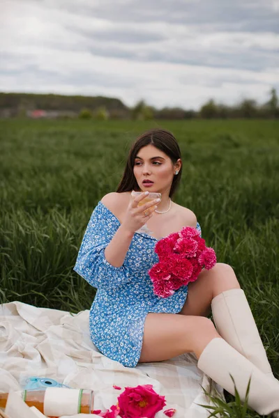 A beautiful young woman in a blue dress with makeup and pink flowers drinks wine at a picnic in nature.