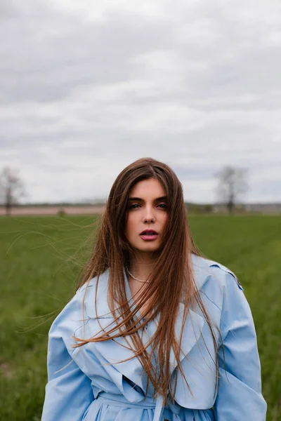 Stylish girl with long dark hair posing in a blue trench coat in a green field. Portrait of a young woman in nature.