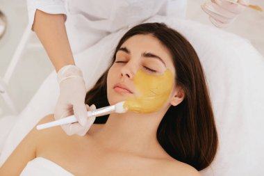 Kozmetik uzmanı kadının yüzüne altın bir maske takıyor. Güzellik salonunda kozmetoloji ve yüz bakımı. Kozmetik işlem. 