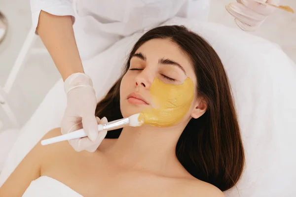 Kozmetik uzmanı kadının yüzüne altın bir maske takıyor. Güzellik salonunda kozmetoloji ve yüz bakımı. Kozmetik işlem. 