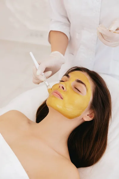 The cosmetologist applies a facial gold mask to the woman's face. Cosmetology and facial skin care in beauty salon. Cosmetic procedure.