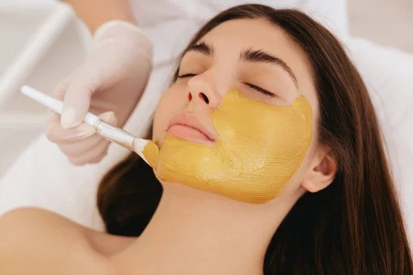 Close-up female face with golden face mask. The cosmetologist applies a facial gold mask to the woman\'s face.  Facial skin care in beauty salon.