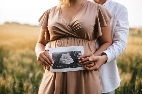 Pregnant woman with husband holding ultrasound baby image. Close-up photo of ultrasound image on the background of a pregnant belly in the hands of mother and father. The concept of pregnancy.