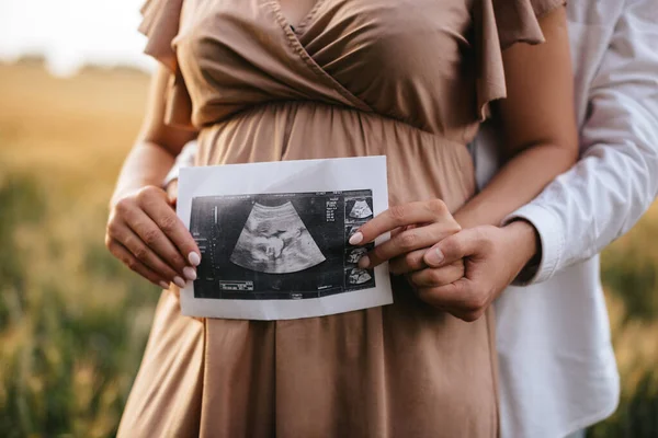 Pregnant woman with husband holding ultrasound baby image. Close-up photo of ultrasound image on the background of a pregnant belly in the hands of mother and father. The concept of pregnancy.