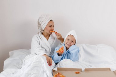 Mom and her little daughter in terry bathrobes and white towels on their heads eat pizza in bed with white sheets. 