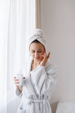 Happy woman in a white bathrobe and with a towel on her head drinks coffee near the window. Lifestyle concept. 