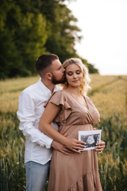 Pregnant woman with husband holding ultrasound baby image. Loving man hugging his pregnant wife from behind in the field. The concept of pregnancy.