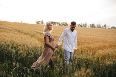 A pregnant woman in a beautiful long dress walks through the field with her husband in a wheat field at sunset. Future parents.