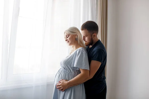 A man hugs his pregnant wife from behind and touches his belly near the window of the house. Pregnant couple. Lifestyle. 