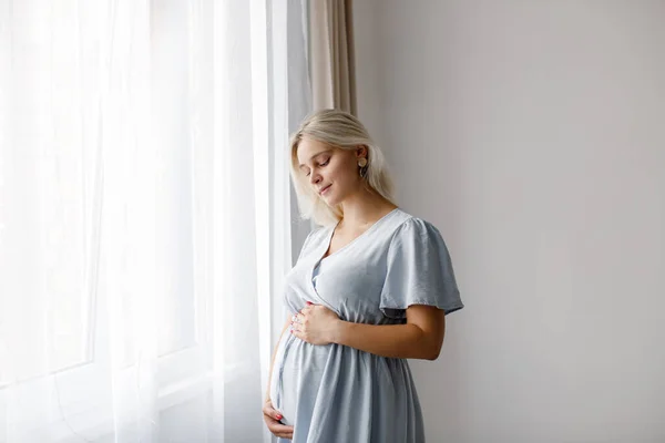 A blonde pregnant woman in a blue dress stands at the window and touches her belly. The pregnancy concept.