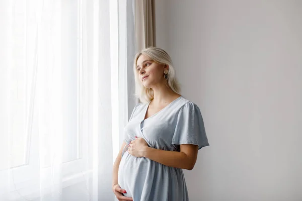 A blonde pregnant woman in a blue dress stands at the window and touches her belly. The pregnancy concept. 