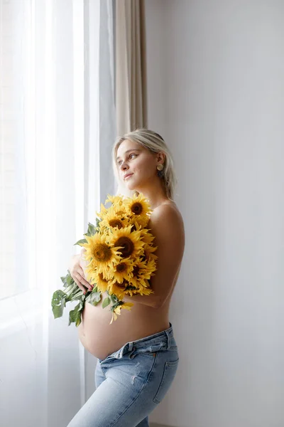 A pregnant woman with a naked belly and jeans covers her body with a bouquet of sunflowers near the window of the house. The concept of pregnancy.