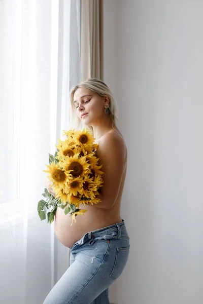 A pregnant woman with a naked belly and jeans covers her body with a bouquet of sunflowers near the window of the house. The concept of pregnancy.