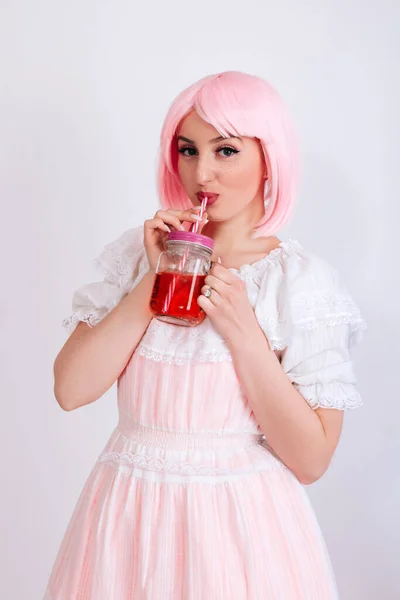 A girl with pink hair and freckles drinks a red drink through a straw on a white background.