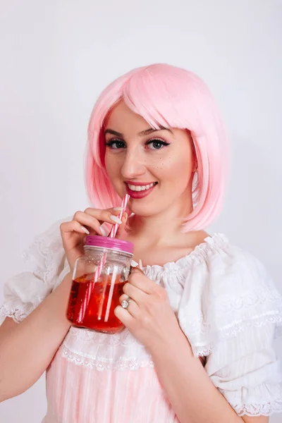 A girl with pink hair and freckles drinks a red drink through a straw on a white background.