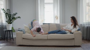 Genç Anne ve Oğul. Genç bir kadın dizüstü bilgisayarla koltukta yatıyor. Açık Renklerle Güzel İç Görünüm.