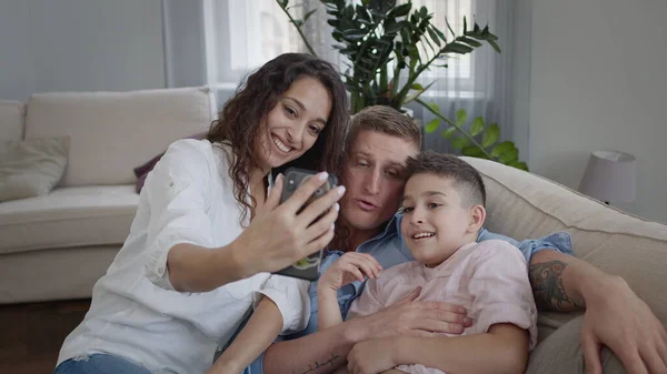 Unge mor tar bilder av sin familie på telefonen. Mamma, pappa og sønn på sofaen. De viser rare ansiktsuttrykk. Familie som har det gøy. – stockfoto
