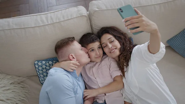 Unge mor tar bilder av sin familie på telefonen. Mamma, pappa og sønn på sofaen. Familie som har det gøy. – stockfoto