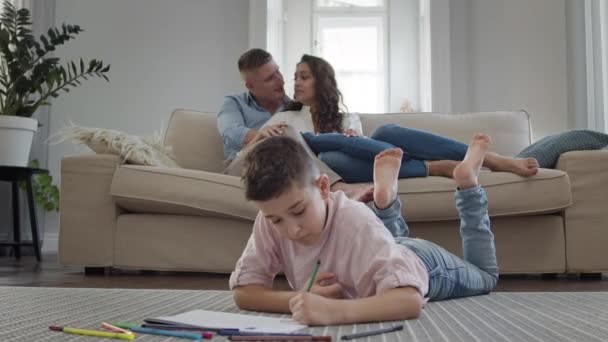 Pojken ligger på golvet i rummet och ritar på papper. På soffan Unga föräldrar. — Stockvideo