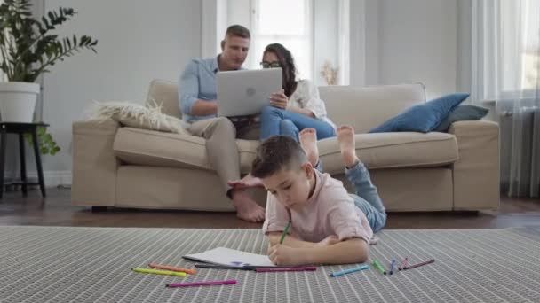 Garçon repose sur le sol dans la chambre et dessine sur un morceau de papier. Sur le canapé Jeunes parents. — Video