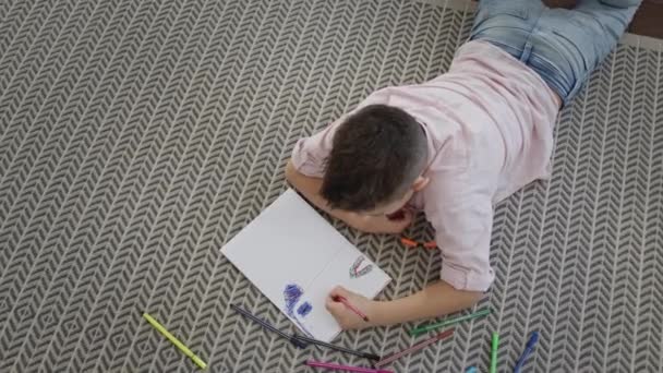 Jongen ligt op de vloer in de kamer en tekent op stuk papier. — Stockvideo